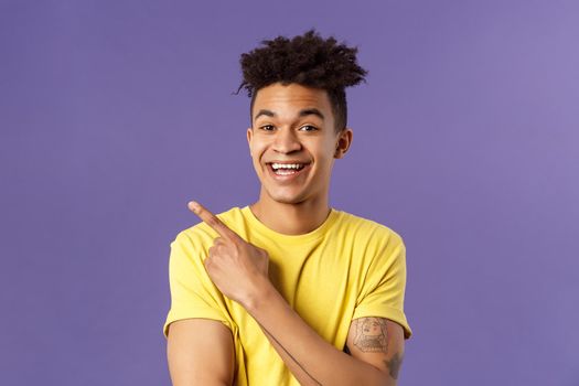Close-up portrait of cheerful, attractive young male student showing promo, pointing at upper left corner, smiling pleased, recommend click link, visit page or download app, purple background.