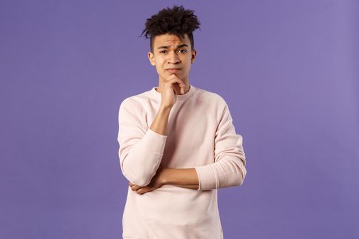 Thoughtful, skeptical young man look judgemental and hesitant at camera while pondering between choices, consider what to do, thinking over purple background, solving troublesome situation.
