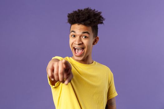 Close-up portrait of upbeat, amazed hispanic man with dreads, young student pointing finger at camera and laughing, recognize someone familiar, standing purple background.