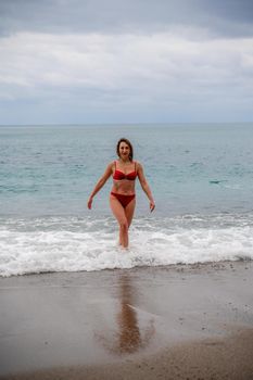 A middle-aged woman with a good figure in a red swimsuit on a pebble beach, running along the shore in the foam of the waves.
