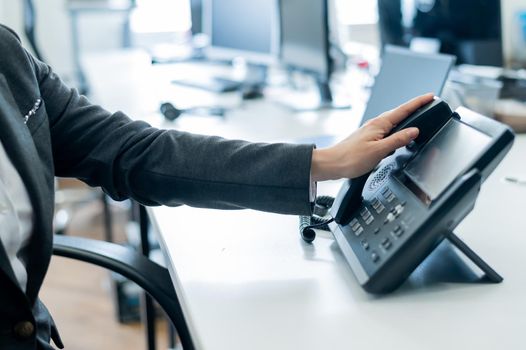 Closeup female hand on landline phone in office. Faceless woman in a suit works as a receptionist answering the phone to customer calls