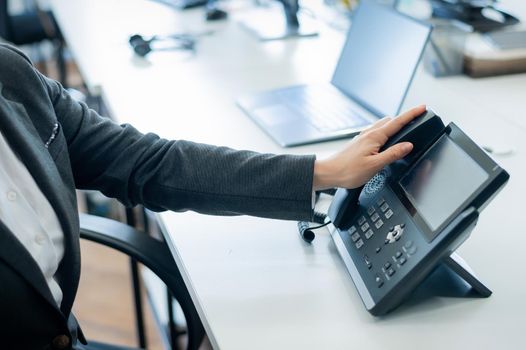 Closeup female hand on landline phone in office. Faceless woman in a suit works as a receptionist answering the phone to customer calls