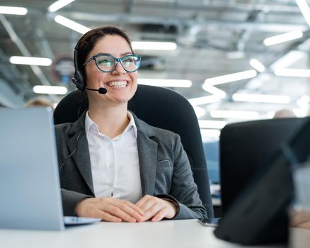 A happy female support operator is sitting at a desk and answering calls. Beautiful smiling woman talking to customers on a headset. Office employee in headphones