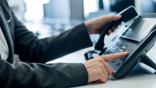 Closeup female hand on landline phone in office. Faceless woman in a suit works as a receptionist answering the phone to customer calls