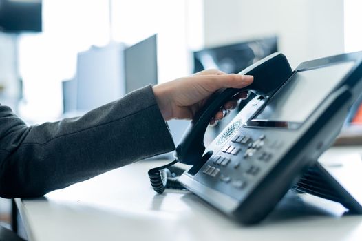 Closeup female hand on landline phone in office. Faceless woman in a suit works as a receptionist answering the phone to customer calls