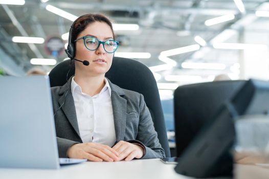 A happy female support operator is sitting at a desk and answering calls. Beautiful smiling woman talking to customers on a headset. Office employee in headphones