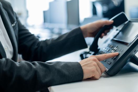 Closeup female hand on landline phone in office. Faceless woman in a suit works as a receptionist answering the phone to customer calls