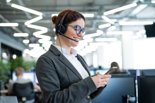 Beautiful caucasian woman in headset is holding a mobile phone while standing in open space office. Friendly female helpdesk operator browsing the screen of a smartphone in the workplace