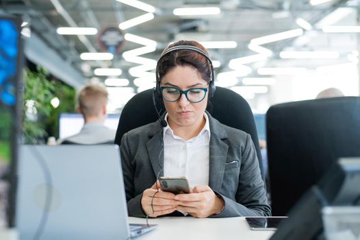 Business woman dressed in a headset is bored and uses a smartphone while sitting at a desk. Female manager is distracted from work by phone.