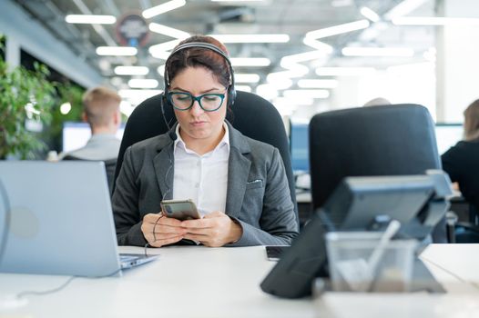 Business woman dressed in a headset is bored and uses a smartphone while sitting at a desk. Female manager is distracted from work by phone.