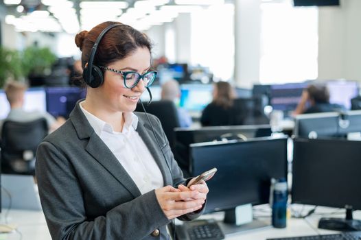 Beautiful caucasian woman in headset is holding a mobile phone while standing in open space office. Friendly female helpdesk operator browsing the screen of a smartphone in the workplace