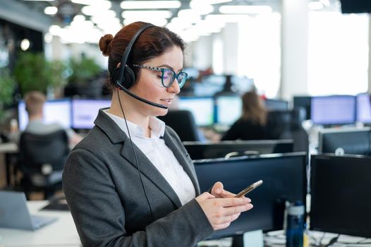 Beautiful caucasian woman in headset is holding a mobile phone while standing in open space office. Friendly female helpdesk operator browsing the screen of a smartphone in the workplace