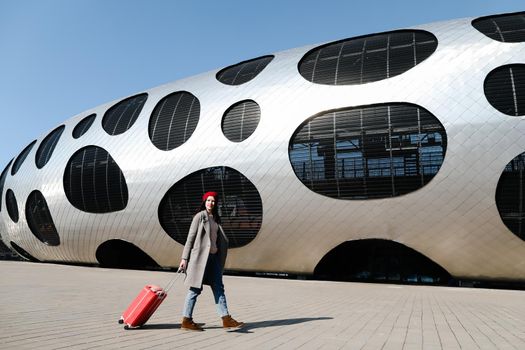 young woman with a red travel suitcase by the futuristic facade of modern architecture. Travel