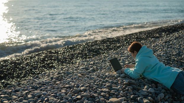 Caucasian woman lying on her stomach on a pebble beach and typing on a laptop. Remote work freelancer