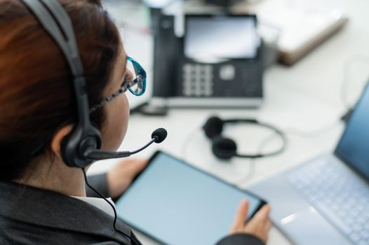 Rear view of a woman in a headset using a digital tablet while sitting at a desk. Friendly female support service operator at work