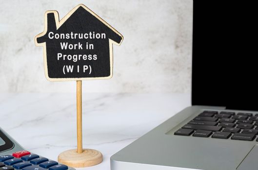 Construction Work in Progress text on wooden house model with notebook and calculator on white table.