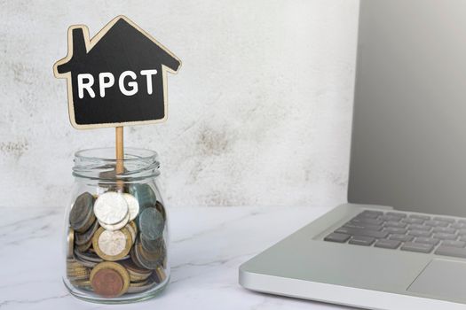 RPGT text on wooden house model with jar full of coins and notebook on white table. Real Property Gains Tax. Property Investment concept.