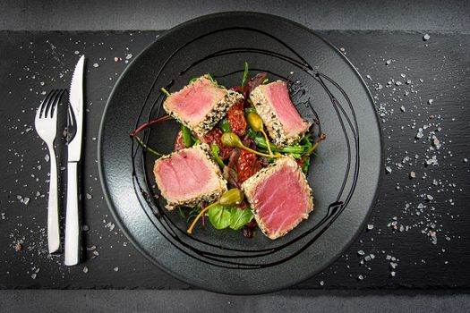 Close up of tuna slices withsesam seeds and fresh vegetables on a dark plate. Top view