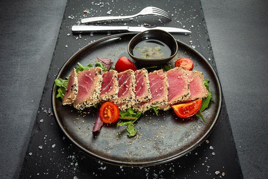 Close up of tuna slices withsesam seeds and fresh vegetables on a dark plate. Top view