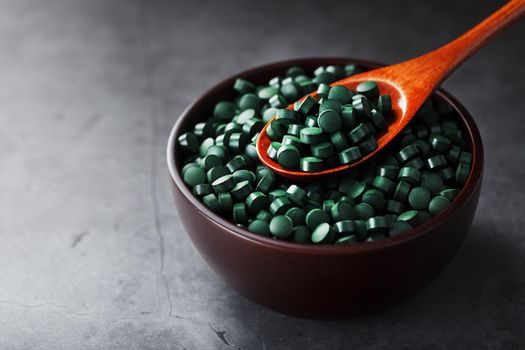 Organic spirulina tablets in a wooden cup with a wooden spoon on a dark background with free space top view