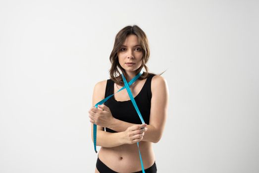 Portrait of a confused young brunette woman in a black underwear holding measuring tape around her neck over white background. Weight loss, slim body, healthy lifestyle concept