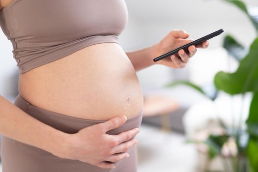 Closeup photo of pregnant female belly. Woman holding and using mobile smart phone app at home interiors. Pregnancy, technology, online shopping, preparation and expectation concept.
