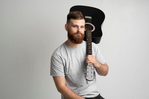 Handsome brunette bearded man musician in a grey t-shirt sitting on a chair and holding a acoustic guitar on a shoulder on a white background studio