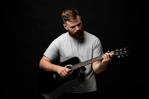 Handsome brunette bearded guitarist plays an acoustic guitar in a black room. The concept of music recording, rehearsal or live performance