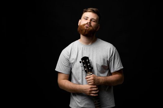 Portraite of handsome brunette bearded man musician, guitarist standing and holding a acoustic guitar in a hand and looks in a camera on a black background studio. Ready to play a music