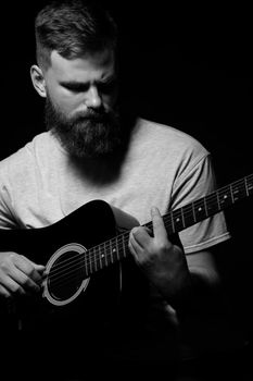 Bearded brutal guitarist plays an acoustic guitar in a black room. The concept of music recording, rehearsal or live performance