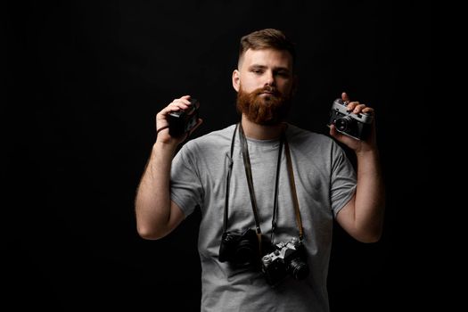 Professional handsome bearded male photographer with bunch of vintage photo cameras in photo studio, isolated on black background