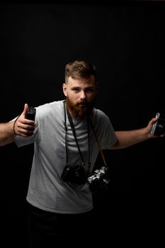Handsome bearded photographer in a grey t-shirt with a bunch of different cameras in a hands and on a shoulder looking on a camera