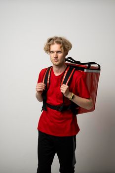 Fast Food Delivery Service Concept. Portrait of smiling male courier wearing red uniform and holding thermal backpack bag isolated on studio background