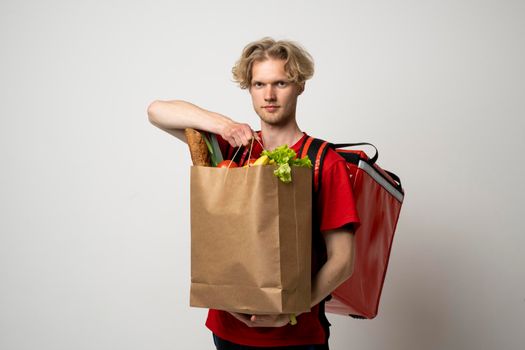 Portrait of pleased delivery man in red uniform smiling while carrying paper bag with food products isolated over white background