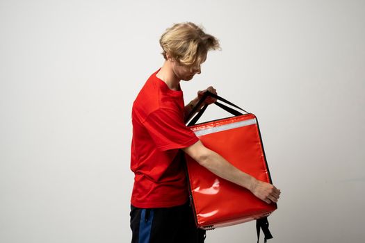 Young courier opening thermal bag on white background. Food delivery service. Delivery guy in a red t-shirt uniform work as courier and holds red thermal food backpack. Service concept