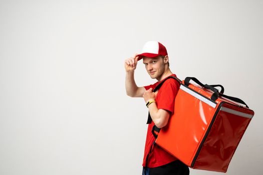 Young courier with thermal bag on color background, space for text. Food delivery service. Delivery guy in a red t-shirt uniform work as courier and holds red thermal food backpack. Service concept