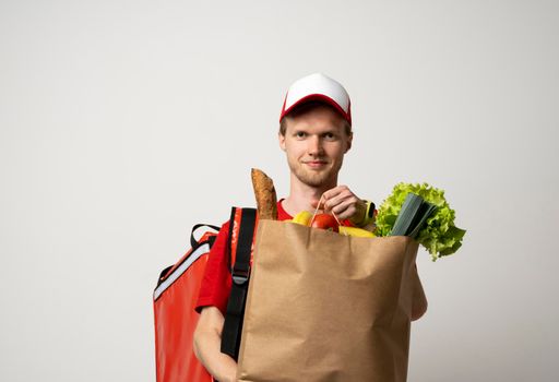 Happy delivery man in red uniform with cap and thermal bag backpack holds brown craft paper bag with food products isolated on gray background studio. Delivery service from shop restaurant to home