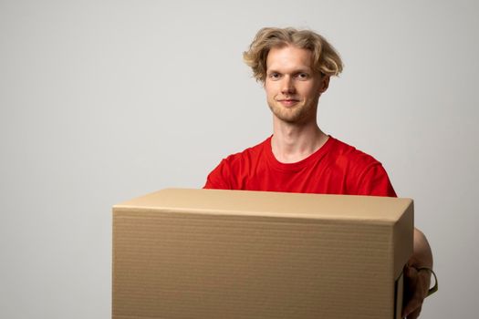 Delivery service. Young smiling courier holding cardboard box. Happy young delivery man in red t-shirt standing with parcel isolated on white background