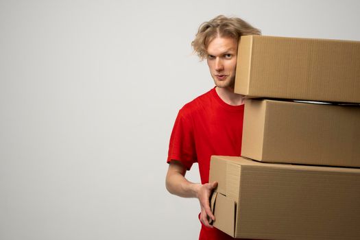 Young delivery man in a red uniform holding a stack of cardboard boxes. Courier delivering postal packages, parcels over white studio background