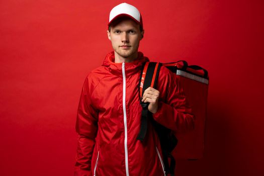 Young courier in a red uniform t-shirt and with red food thermo bag on a shoulder standing isolated on red background studio. Food delivery service