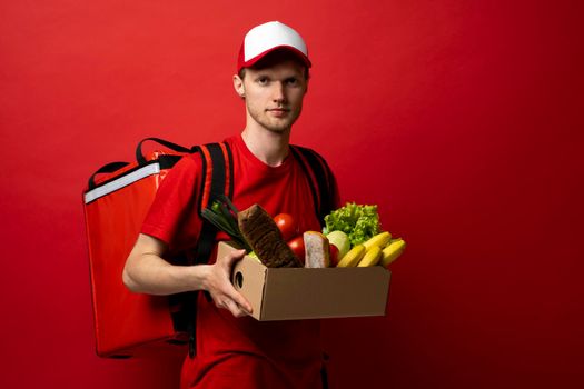 Delivery man in red uniform holds craft cardboard box with food isolated on white background, studio portrait. Service concept