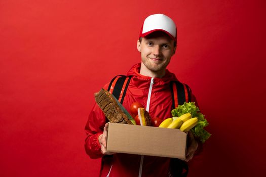 Young delivery man carrying paper box bag of grocery food and drink from store isolated on red studio background. Copy Space. Delivery Concept