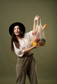 Eco friendly shopping. Smiling young woman holding string reusable mesh bag with organic fruits and vegetables. Positive female standing on green studio background