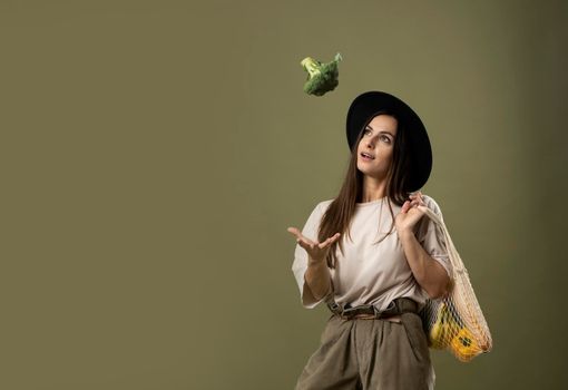 Smiling brunette young woman in a beige t-shirt and a hat holding a reusable mesh bag with fresh vegetables and fruits on a shoulder and throwing a broccoli in a air. Zero waste concept