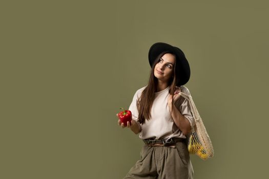 Sustainable lifestyle. Young woman in a black hat holding mesh grocery bag with vegetables on shoulder and red pepper. Zero waste, plastic free. Eco friendly concept