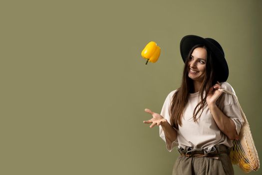 Woman in beige t-shirt throw up a yellow pepper in a air by one hand and holding reusable cotton eco bags on a shoulder