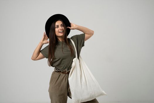 Brunette woman in green t-shirt and black hat holding cotton shopper bag with vegetables, products in white room. Eco friendly shopping bags. Zero waste, plastic free concept