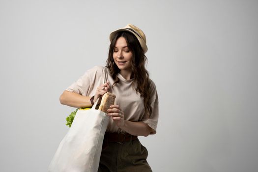 Smiling young woman in beige t-shirt and a hst with a mesh eco bag full of vegetables and on a white studio background. Sustainable lifestyle. Eco friendly concept. Zero waste