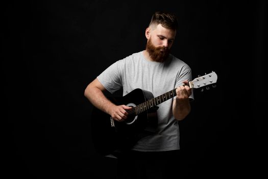 Handsome brunette bearded man musician plays an acoustic guitar in a black room. Ready to play a music