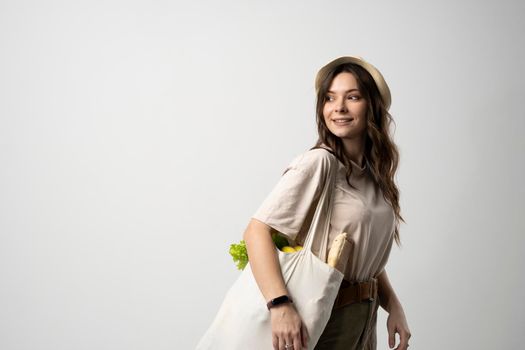 Smiling woman in a t-shirt and a hat holding reusable cotton shopping bag with vegetables, bread and greens. Concept of no plastic, zero waste, plastic free, sustainable lifestyle